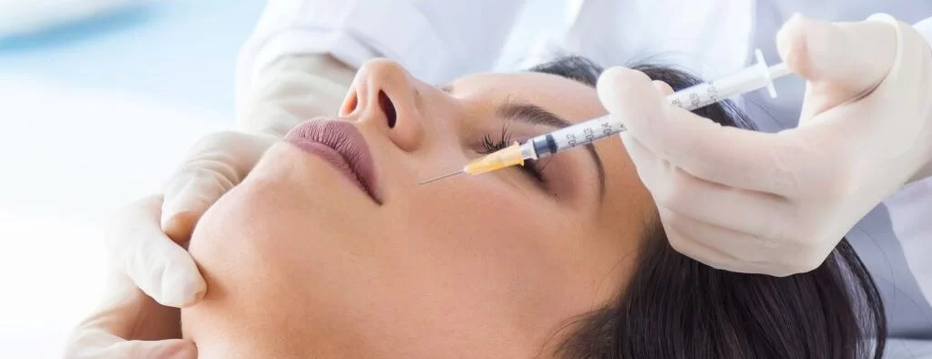 Mujer recibiendo un tratamiento estético con inyección en la cara, enfocándose en la zona de los ojos, en un ambiente clínico.