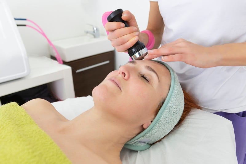 Mujer recibiendo un tratamiento facial en un spa, con una esteticista utilizando un dispositivo de belleza en su frente. La paciente está relajada, con una toalla verde y una banda en la cabeza.