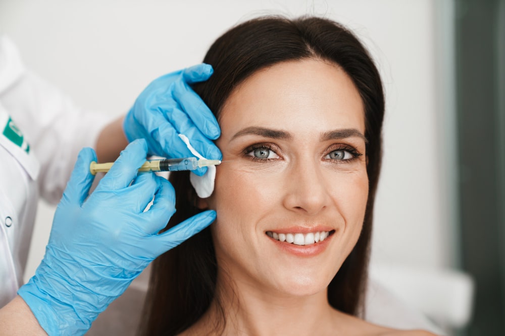 Mujer sonriente recibiendo un tratamiento estético con inyecciones en la zona de los ojos, mientras un profesional de la salud la asiste.